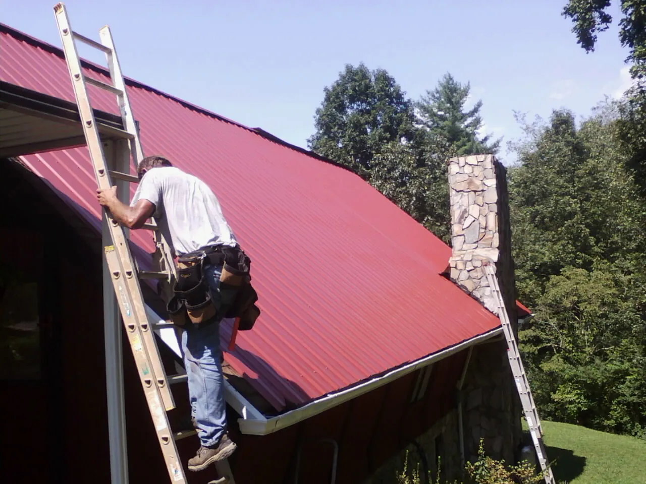 Roofer on a ladder beside a red metal roof and stone chimney — Soffit & Fascia Repair by Roofing Brothers in Tillamook